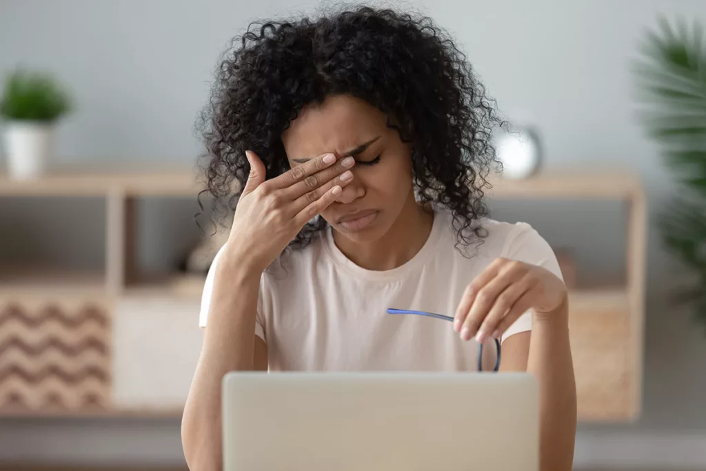 A young woman with a laptop rubs her eyes while experiencing an ocular migraine