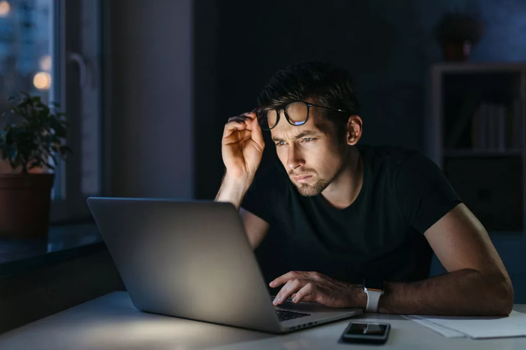 A man sits at his desk, struggling to look at a laptop due to worsened night vision.