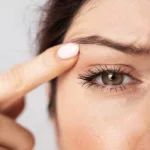 A woman with hazel eyes points a finger to her droopy eyelids